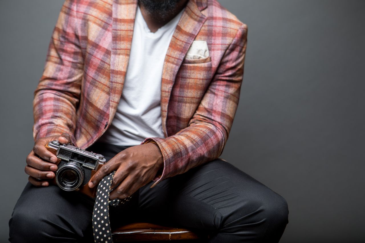 quality-assurance-01 A fashionable man in a plaid blazer holds a vintage camera, seated indoors.