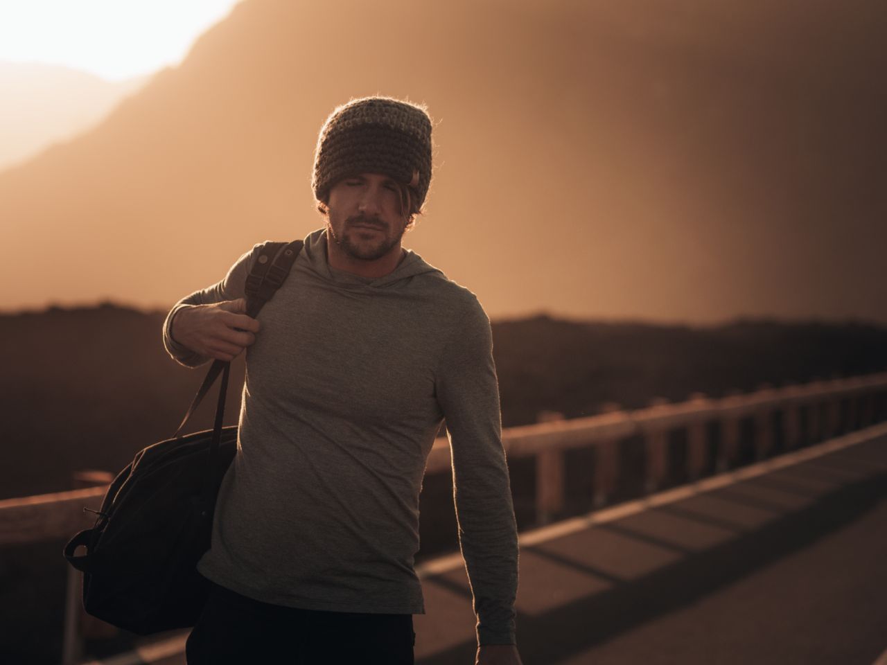 A man in a knit hat and hoodie walks with a bag at sunset on a scenic road in Santa Cruz de Tenerife.