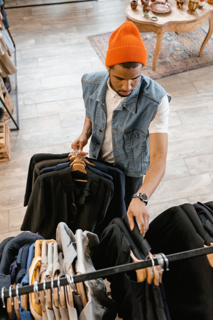 social-feed-img-04 Man examines clothing in boutique wearing denim vest and orange beanie.