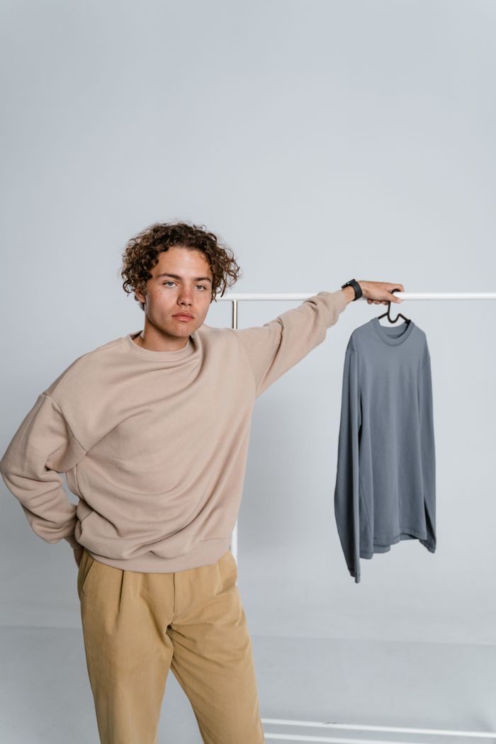 social-feed-img-05 Young man with curly hair posing in a minimalist studio with a sweater on a hanger.