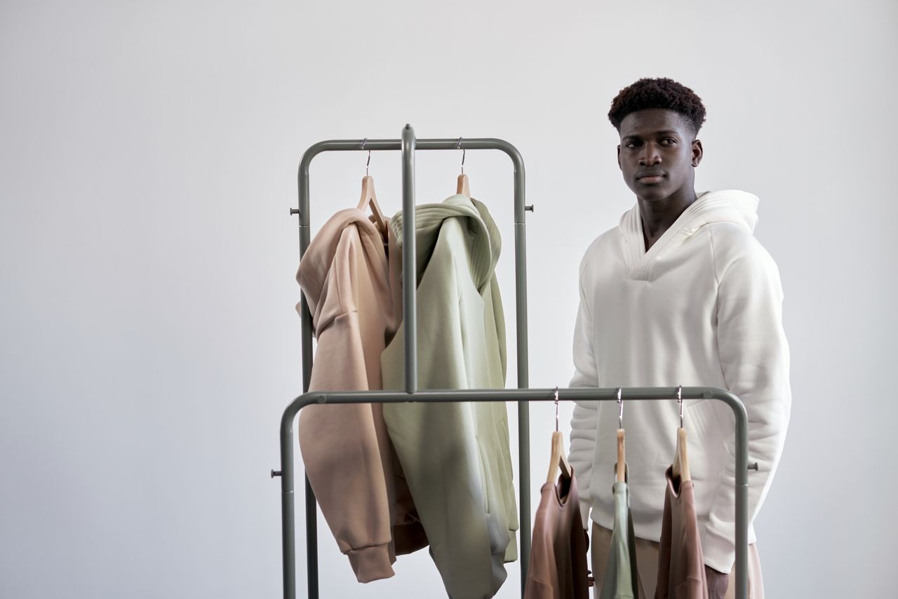social-feed-img-01 Young man stands beside a rack of stylish hoodies in a minimalist setting.